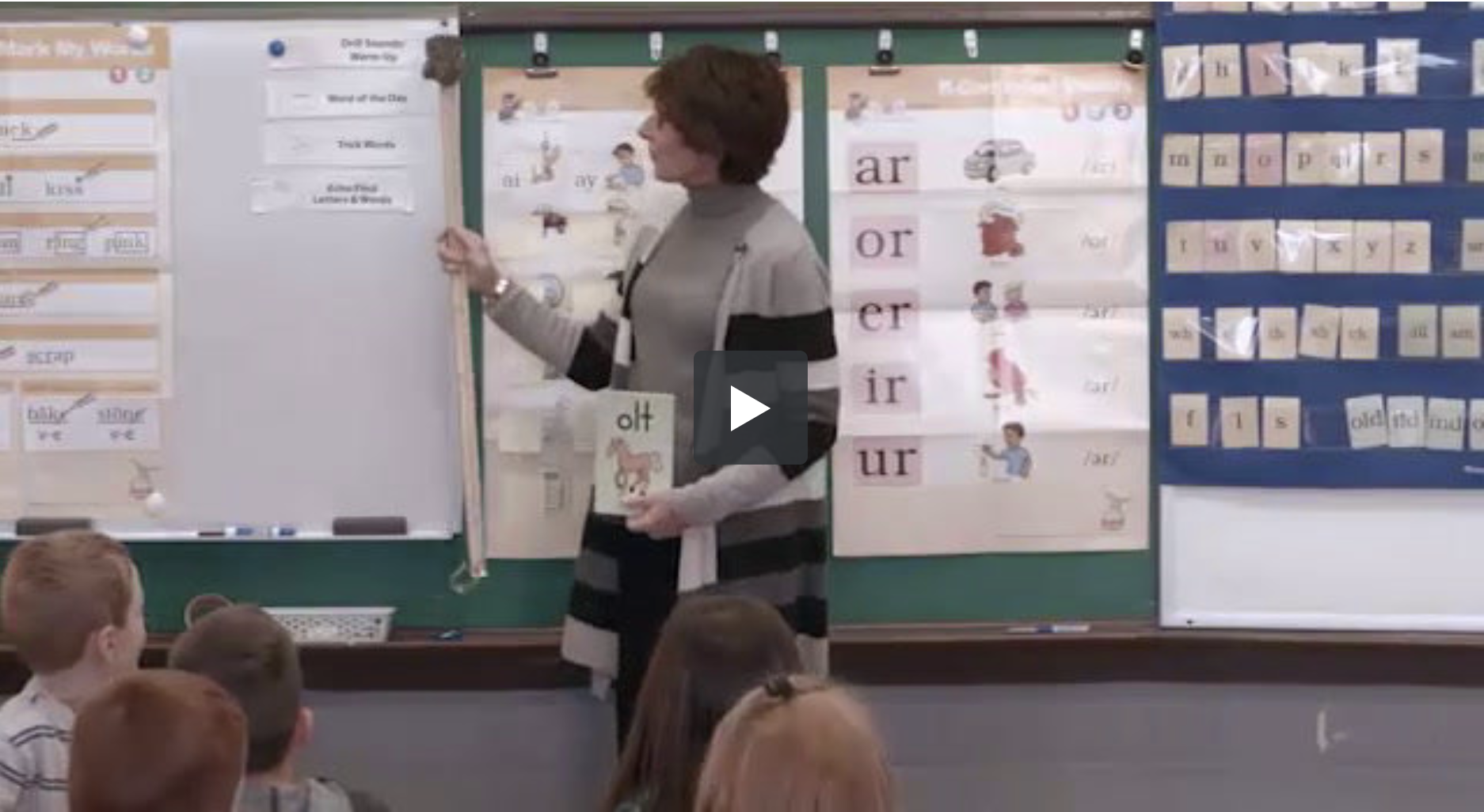 A teacher in a striped sweater is standing at the front of a classroom, pointing to a whiteboard with lesson materials. Several children are seated on the floor facing her. Charts with words and images are displayed on the wall behind her.